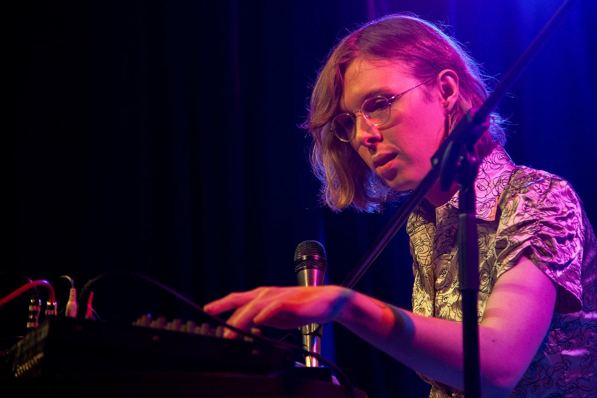 Mira Martin-Gray performing experimental electronic music. She is seated, shot in profile and lit with bisexual lighting. There is a microphone in front of her, and she is touching a mixing board gently. She is marginally attractive, with chin-length blonde hair and round glasses, wearing a short-sleeve gold blouse with floral embroidery.